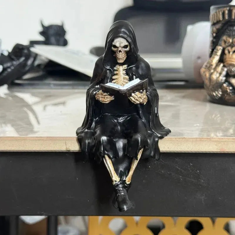A gothic grim reaper figurine seated on a black shelf, holding an open book. The figure is dressed in a glossy black hooded robe with gold-painted bones and skeletal details, set against a blurred home office background.