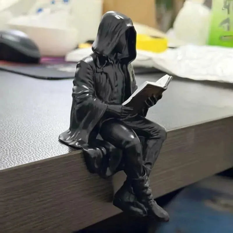 A small black figurine of a hooded seated person reading a book, placed on the edge of a desk in a real indoor setting with blurred office items in the background.