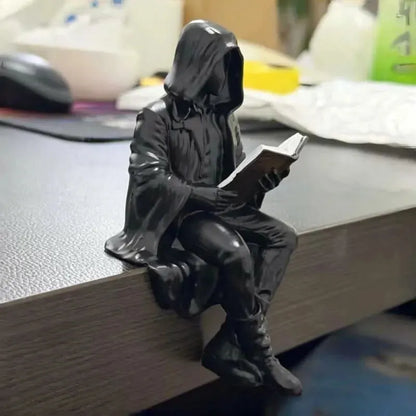 A small black figurine of a hooded seated person reading a book, placed on the edge of a desk in a real indoor setting with blurred office items in the background.
